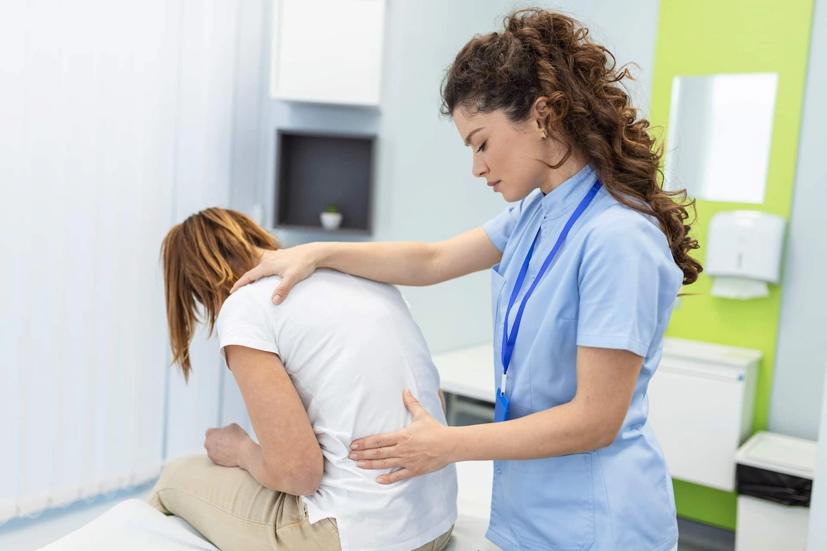 A healthcare professional in blue scrubs places hands on the back of a seated patient with reddish-brown hair, who is wearing a white t-shirt and light brown pants, in a medical room.