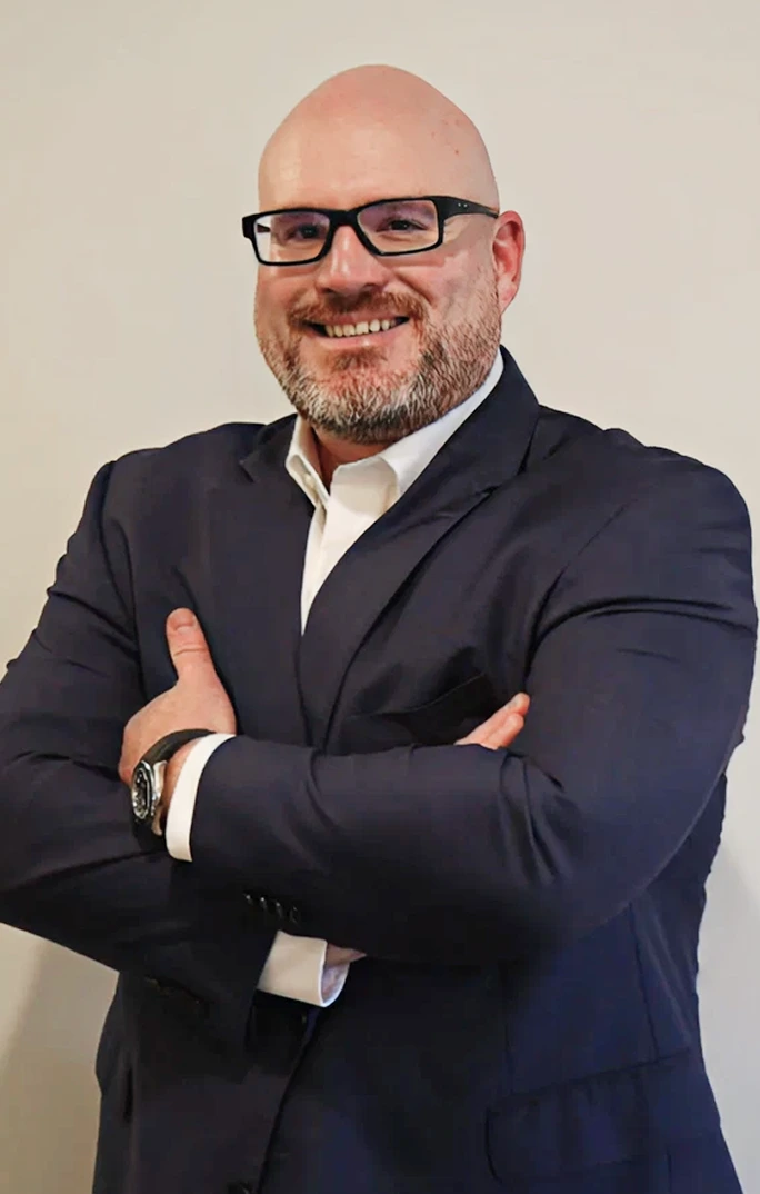 A bald man with a beard and black glasses, wearing a dark suit and white shirt, smiles with his arms crossed.