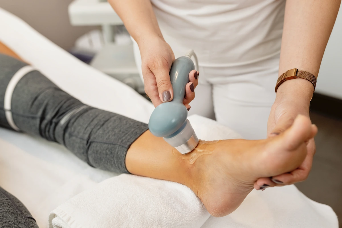 Close-up of a person's ankle receiving treatment with a handheld device and gel, administered by a practitioner.