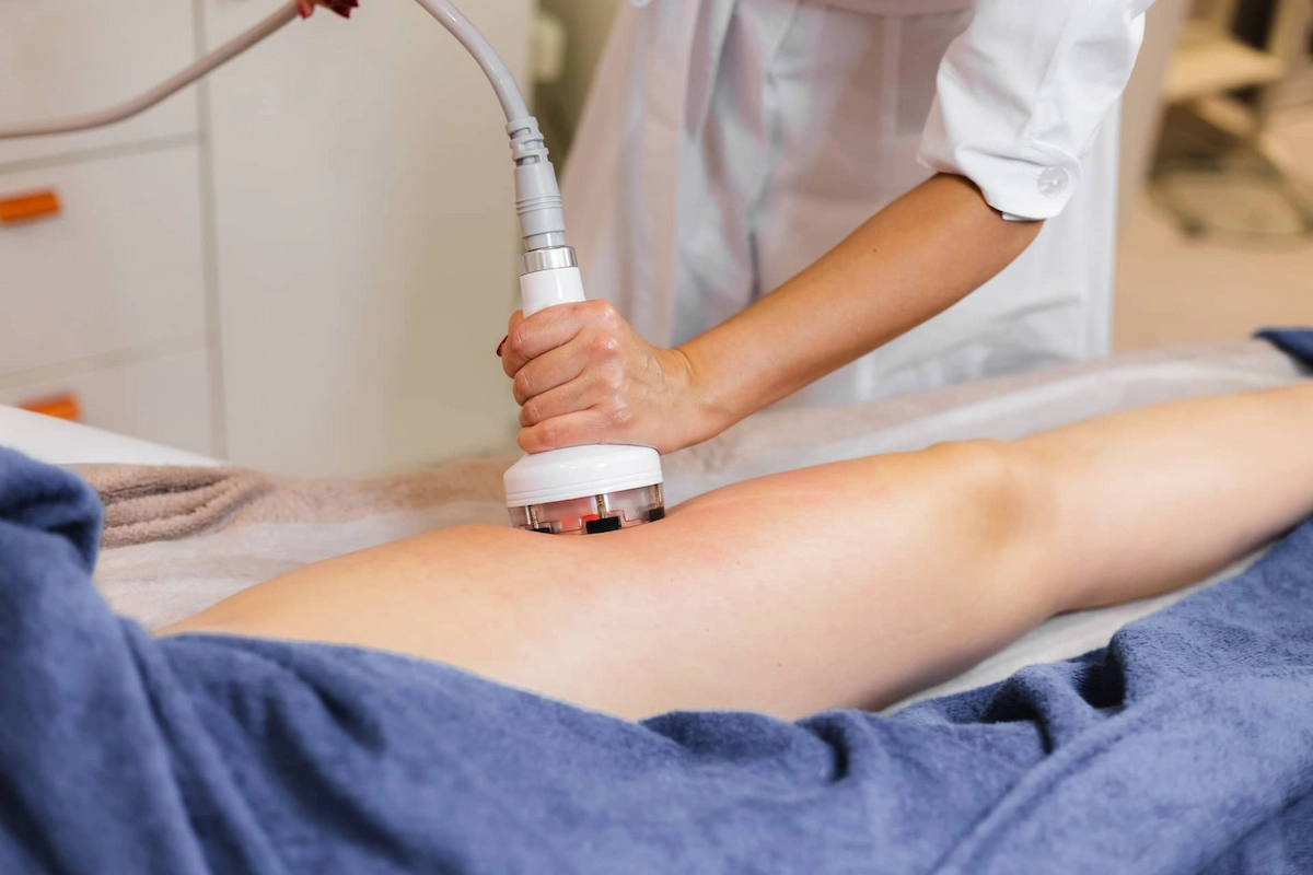 A professional uses a white and grey handheld device with red lights on a client's upper leg, which is partially covered by a blue towel.