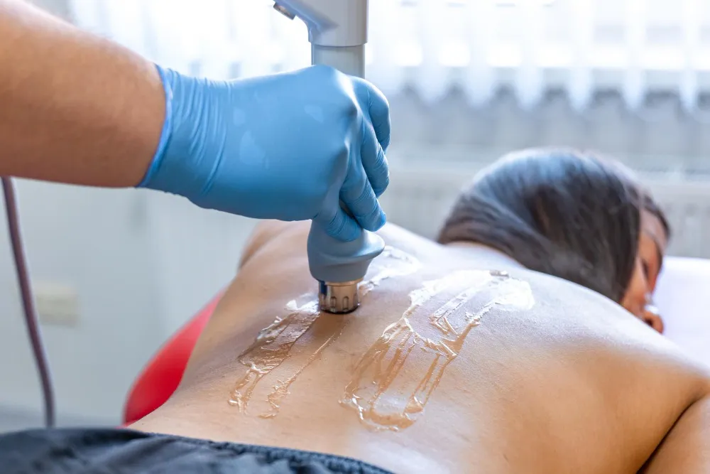 A gloved hand applies a medical device to a person's bare back, which has clear gel applied in streaks.