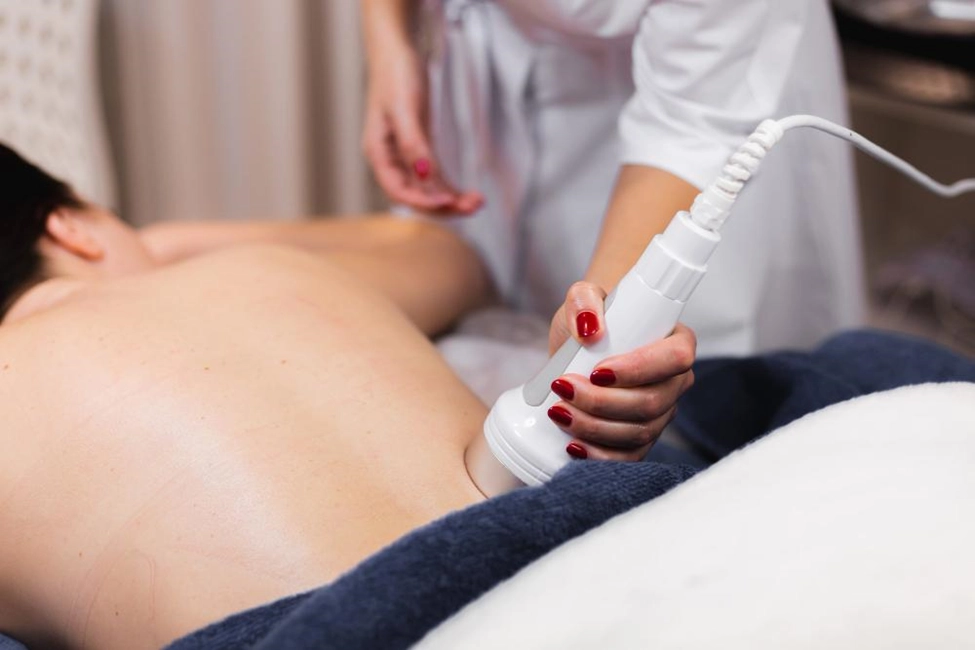 A person lies face down as a therapist uses a white handheld device on their lower back. The therapist's hand has red nail polish.