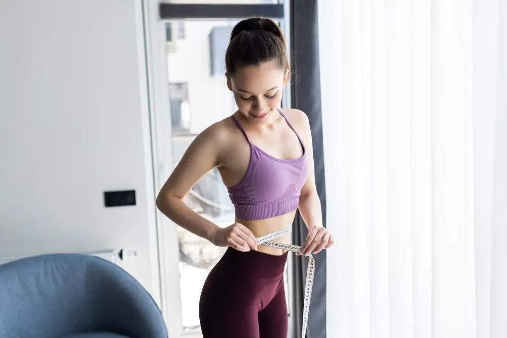 A young woman in a purple sports bra and maroon leggings measures her waist with a tape measure, smiling as she looks down.