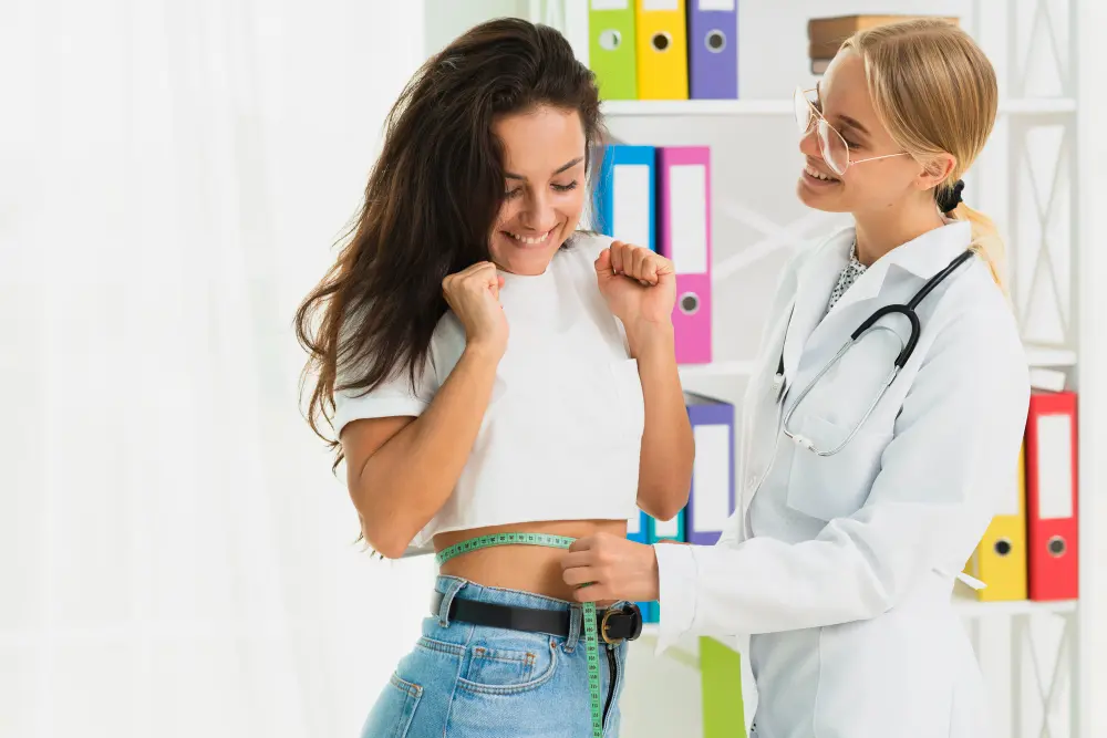A doctor in a white lab coat measures the waist of a smiling woman in a white crop top and jeans with a green tape.