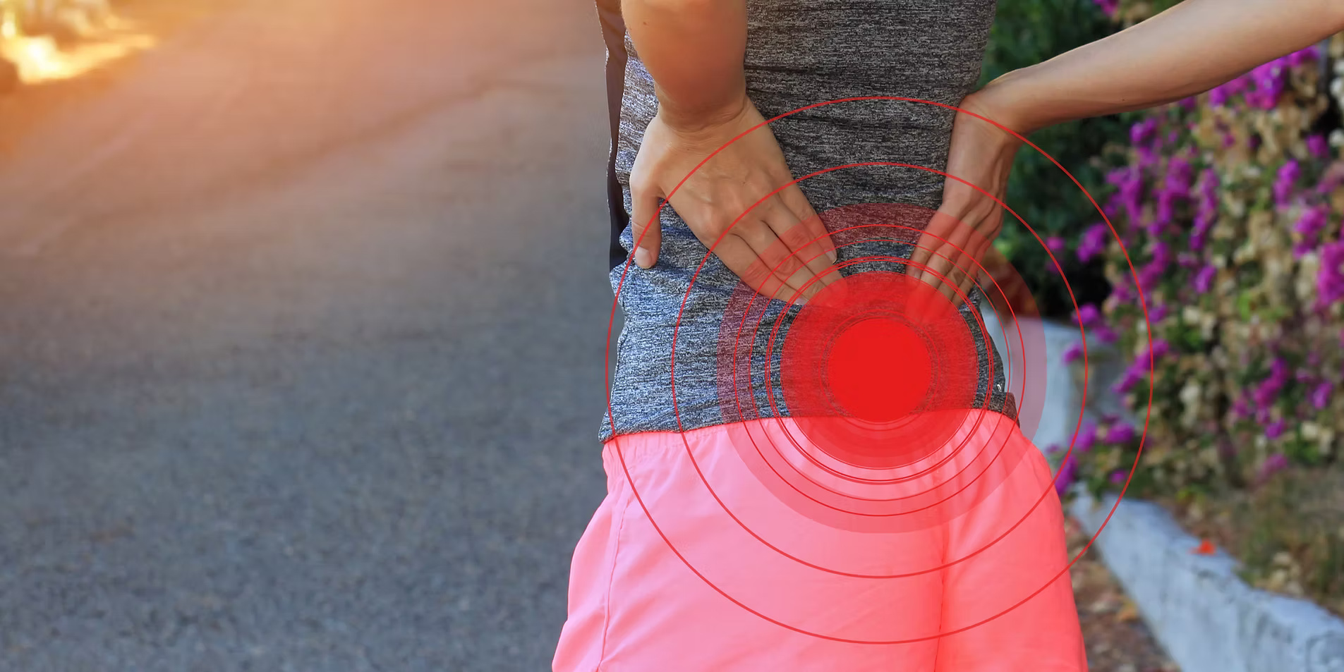 A person from behind, wearing athletic wear, holding their lower back with both hands. Red circular graphics indicate pain in the lower back and hip area. The background is blurred, showing a road and foliage with purple flowers.