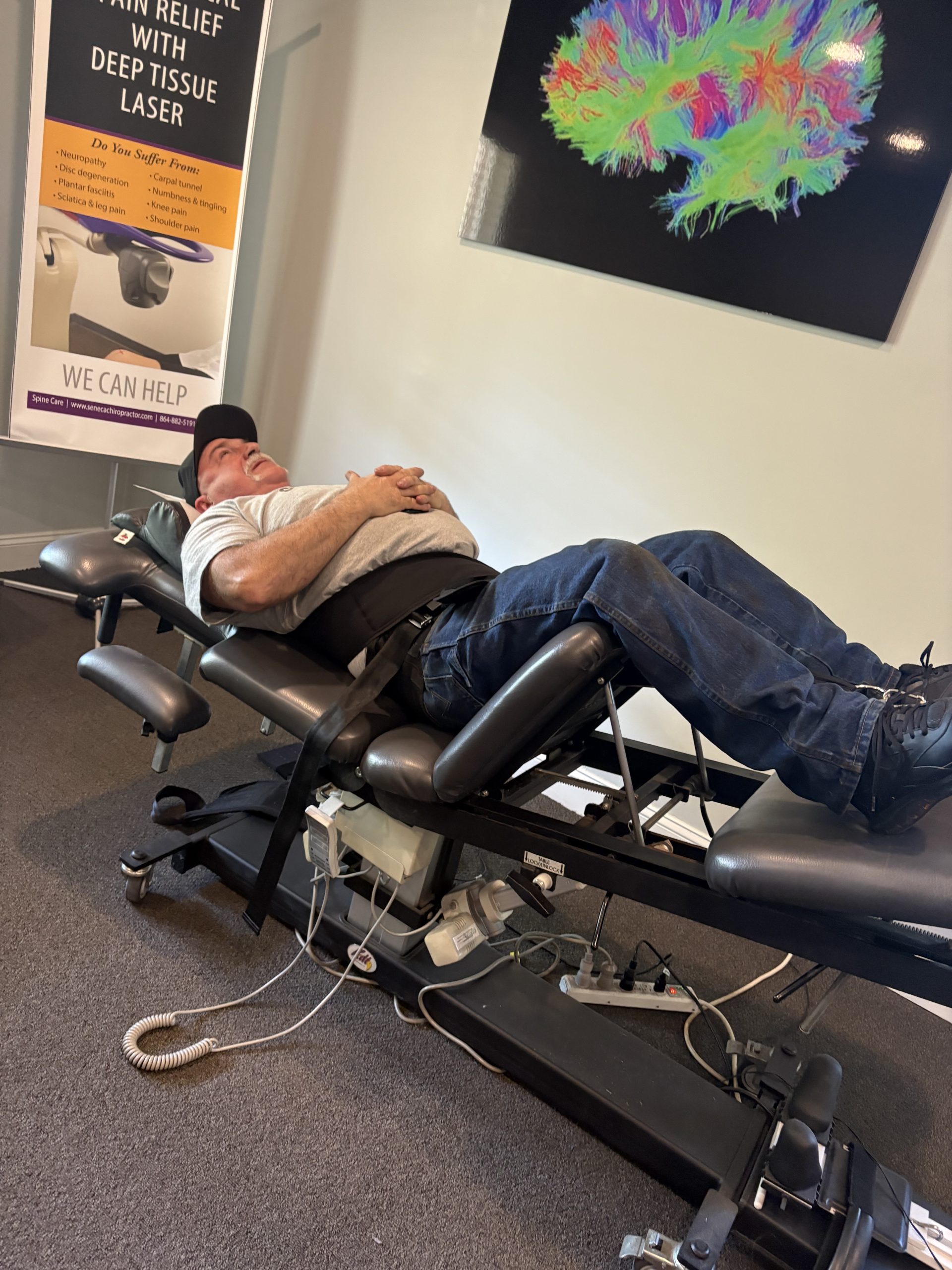 An older man lies on his back on a black medical treatment table, secured by a waist strap. A banner for 'Deep Tissue Laser' and a colorful brain scan image are in the background.
