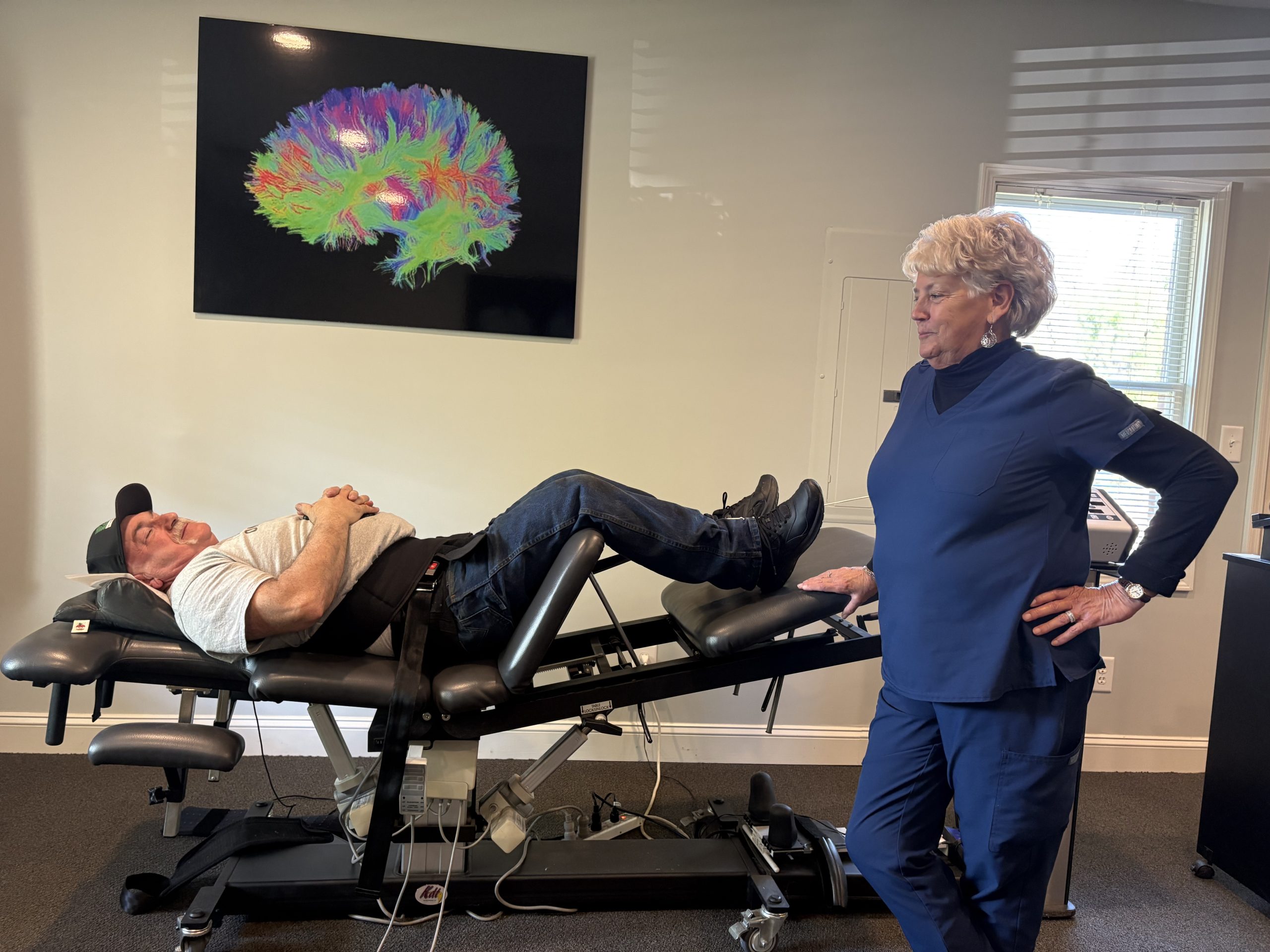 An older man lies on a chiropractic table with a harness, while an older woman in scrubs stands beside him, observing.