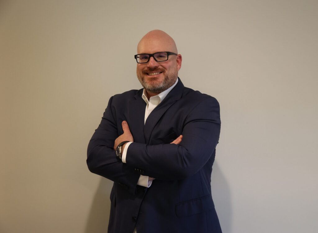 Bald man with beard and glasses wearing a suit, arms crossed, smiling at the camera.