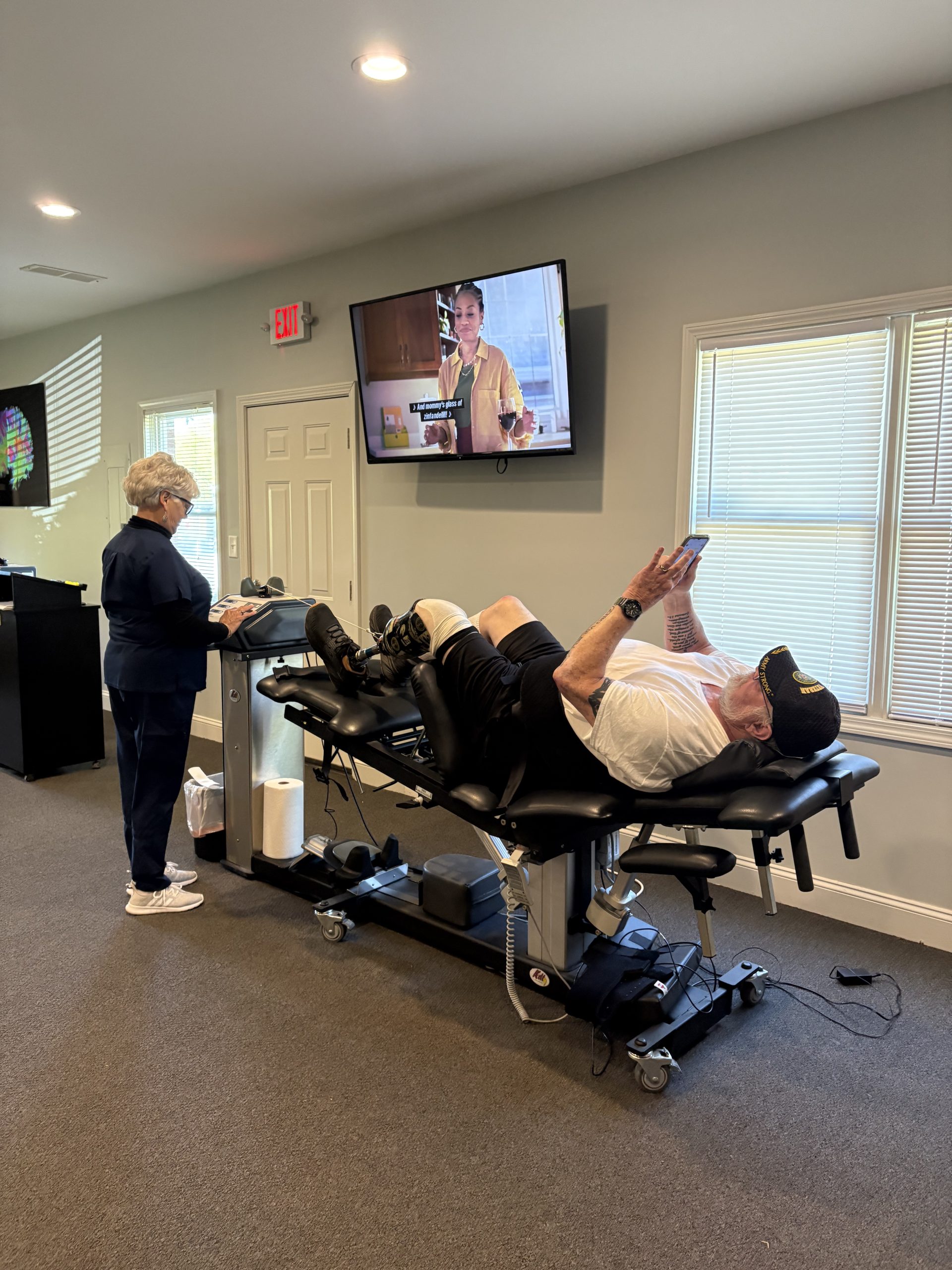 A man with a prosthetic leg lies on a therapy table, looking at his phone, while a woman in scrubs operates a machine next to him.