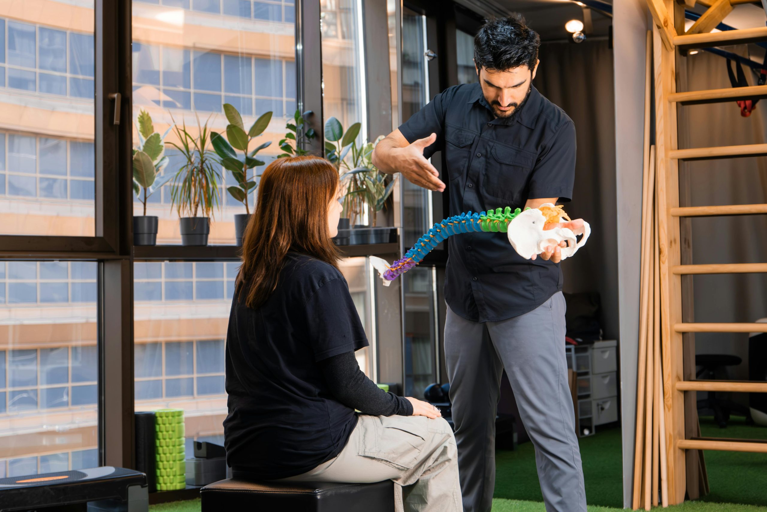 A man in a black shirt holds a colorful spine and pelvis model, explaining it to a woman seated on a stool in a modern clinic with large windows.