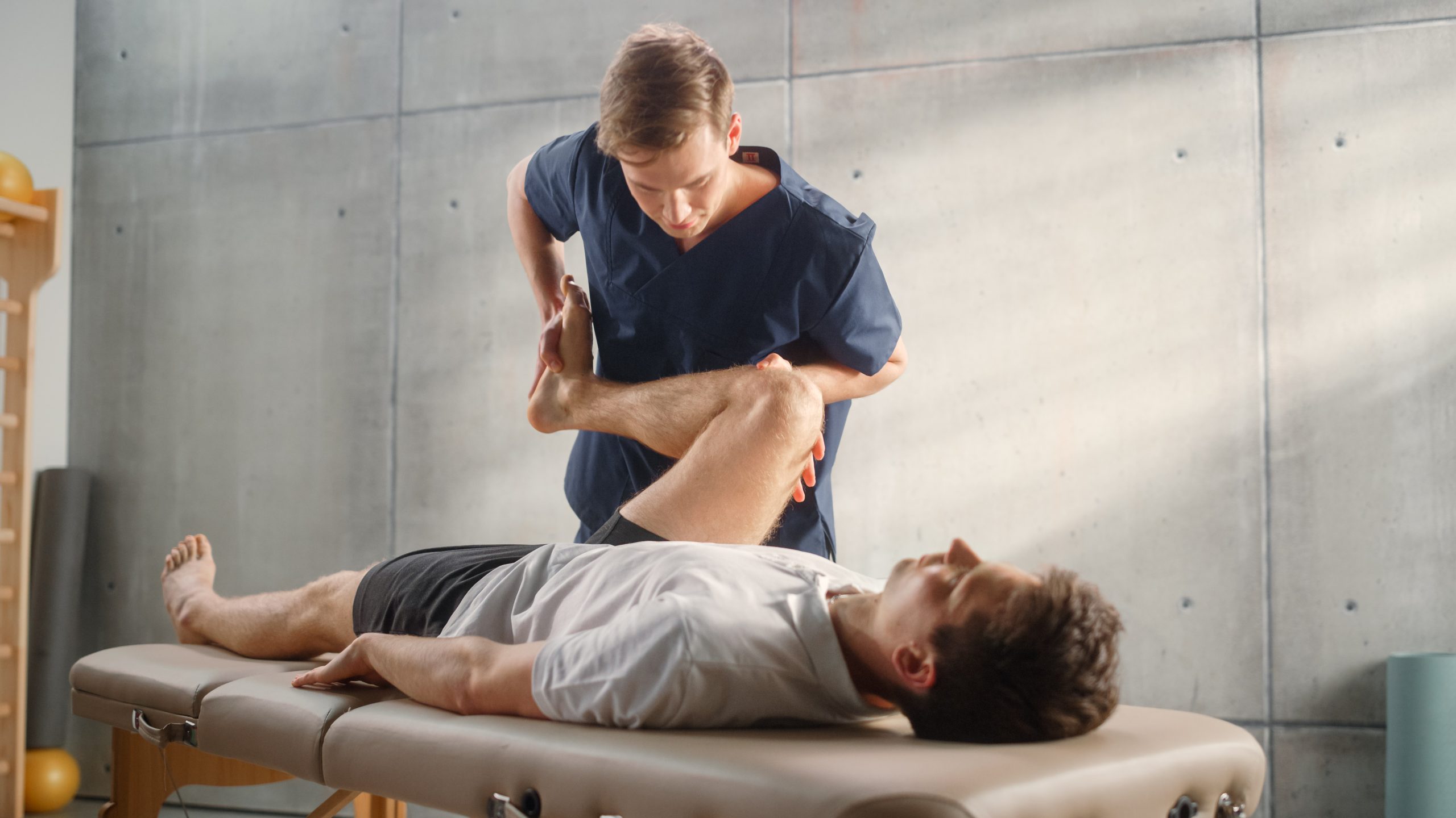 A male physiotherapist manipulates the leg of a male patient lying on an examination table.