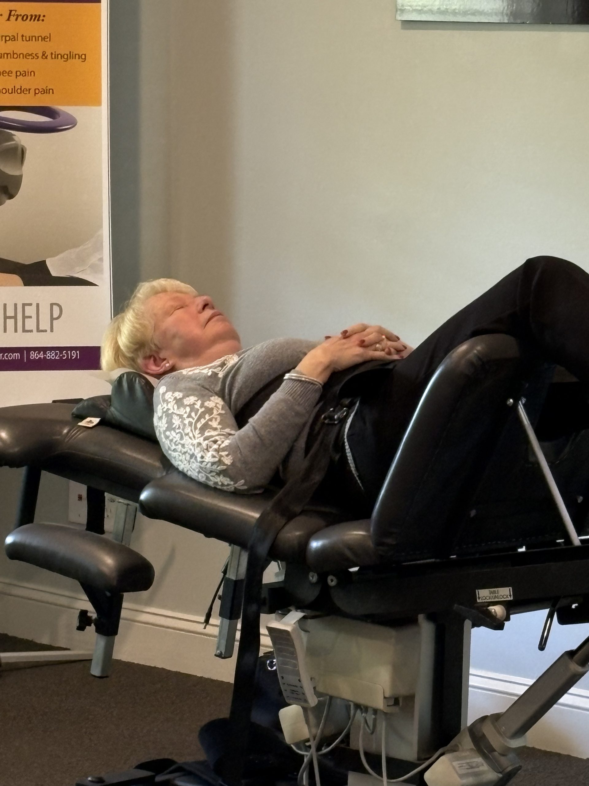An older woman with blonde hair lies on her back on a black therapy table, eyes closed and hands clasped. A poster with medical-related text is partially visible in the background.