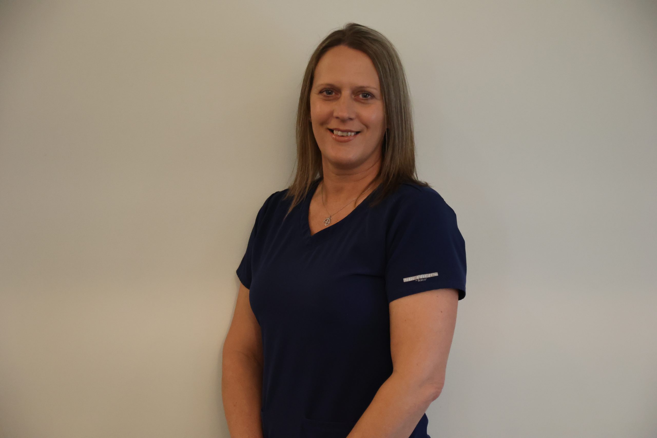 A smiling woman with light brown hair wearing a dark blue scrub top and a silver necklace.