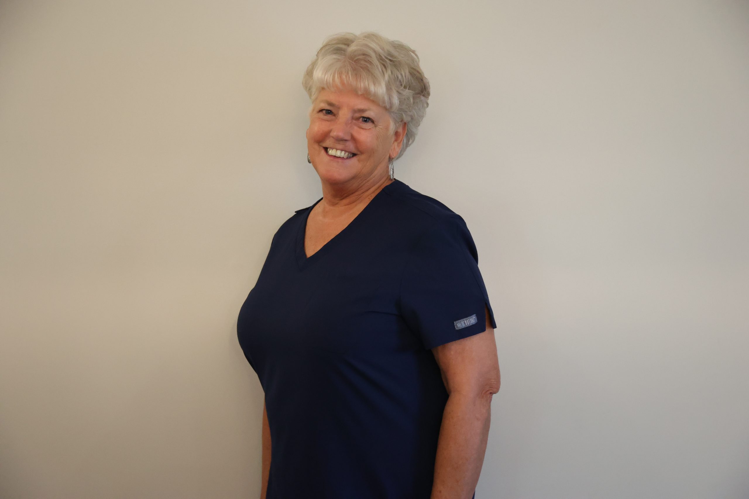 A smiling woman with gray hair in a navy blue scrub top.