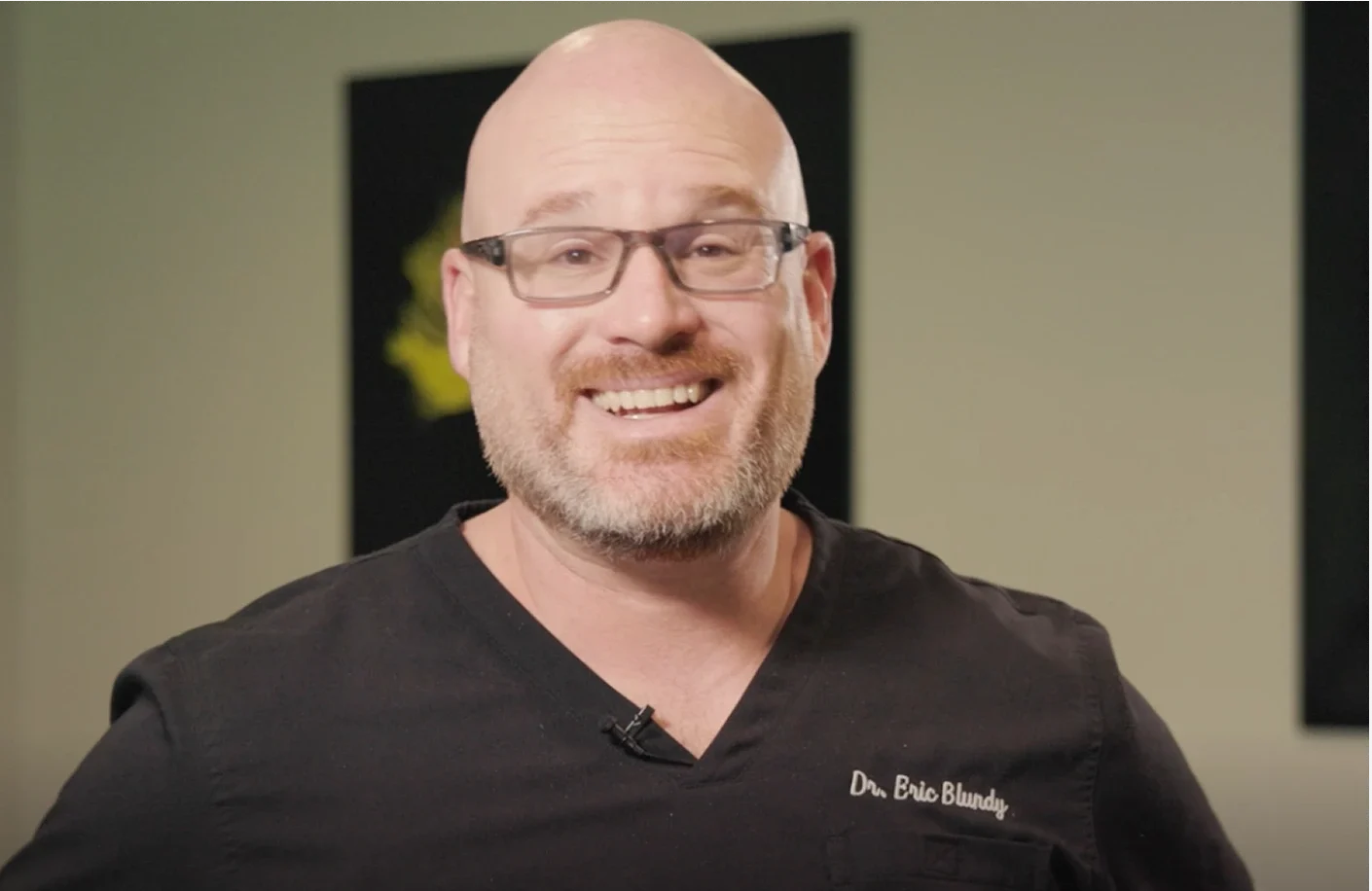 A smiling bald man with a beard and glasses, wearing black scrubs with "Dr. Eric Blundy" embroidered, stands in front of a light wall. A video play button and text 'Watch an Introduction to Spine Care South Carolina' are at the bottom.