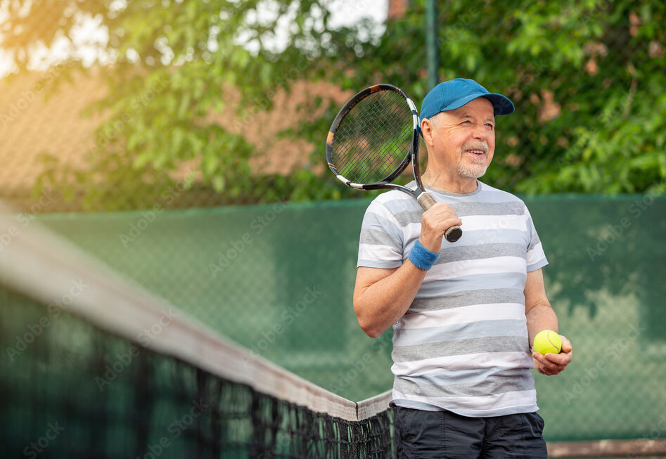 An older man in a blue cap and striped shirt stands on a tennis court, holding a tennis racket over his shoulder and a tennis ball in his hand, smiling.