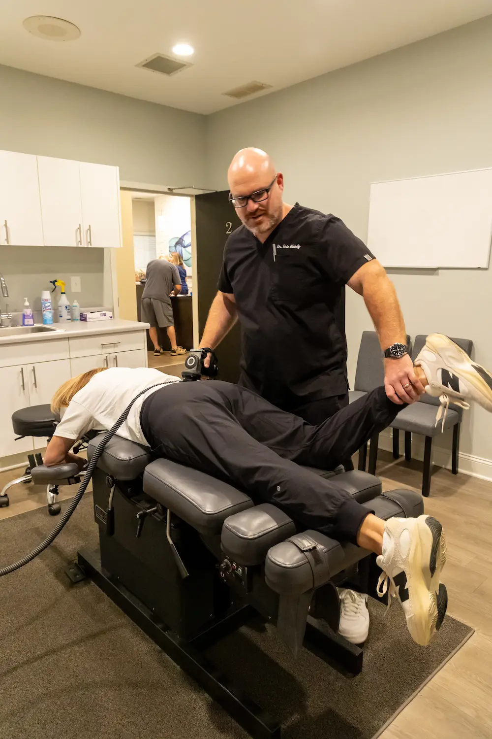 A chiropractor adjusts a patient lying face down on a table, lifting her leg while using a tool on her lower back.