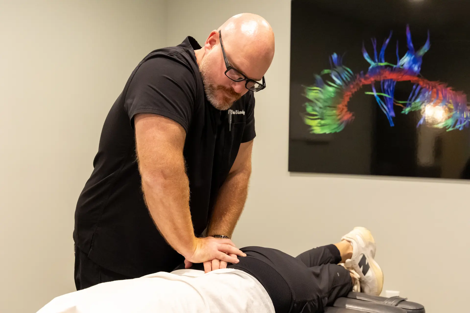 A bald man with glasses and a beard adjusts a patient's lower back on a treatment table. The patient wears black pants and white sneakers. A colorful abstract artwork hangs on the wall behind them.