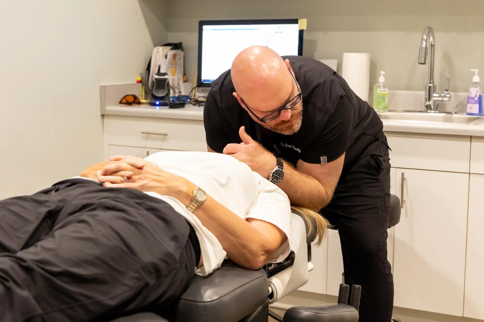 A male chiropractor in black scrubs adjusts a patient lying on a chiropractic table.