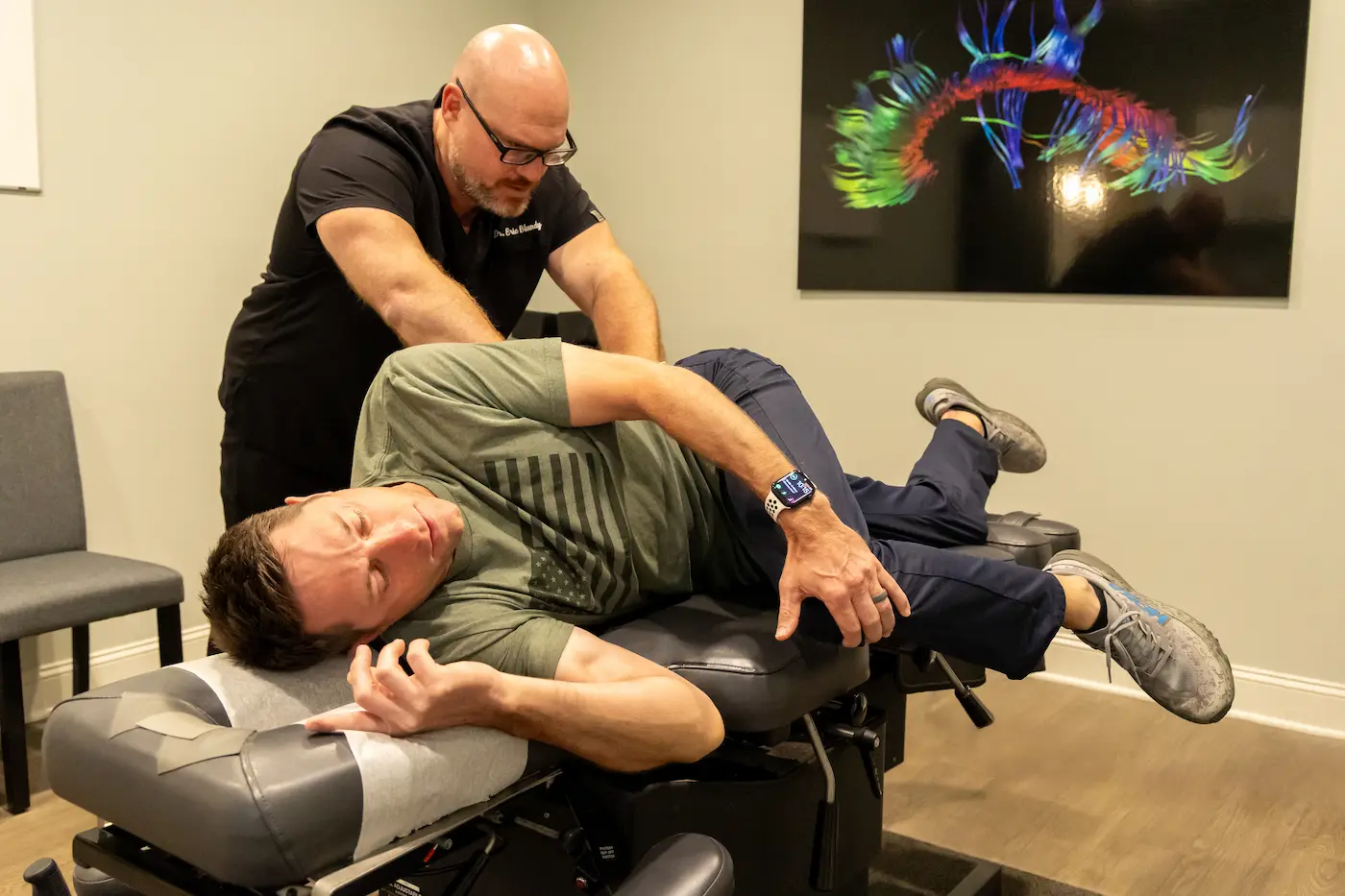 A chiropractor in black scrubs adjusts a male patient lying on his side on a chiropractic table.
