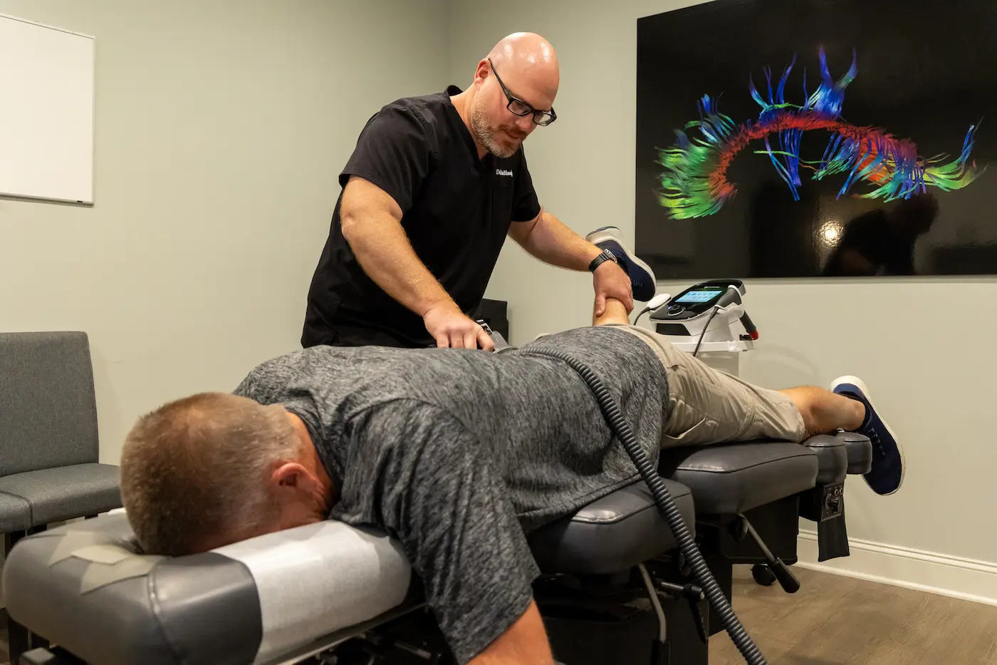 A practitioner treats a patient lying face down on a chiropractic table with a medical device.
