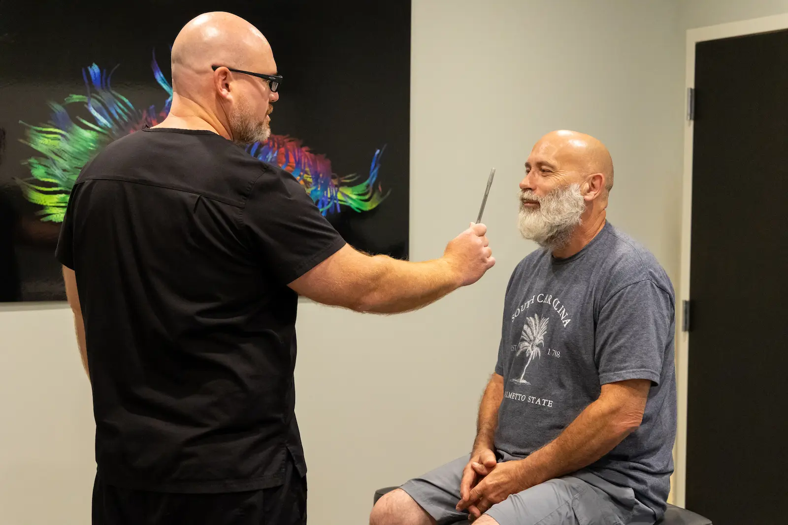 A bald man in black scrubs holds a silver instrument near the face of a seated bald man with a white beard, who is wearing a gray t-shirt.