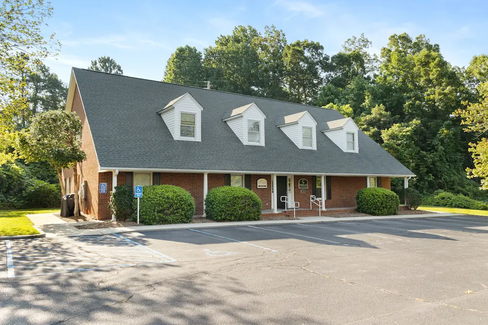 A red brick building with a dark gray roof and four dormer windows, featuring accessible parking spaces in the foreground and surrounded by green trees and bushes under a blue sky.