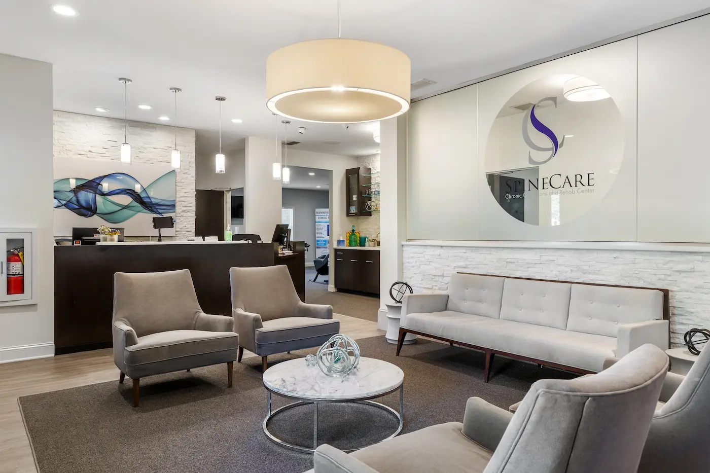A modern and clean reception area with grey armchairs, a matching sofa, a marble coffee table, and a dark wood reception desk. A 'SpineCARE' logo is visible on a frosted glass partition.