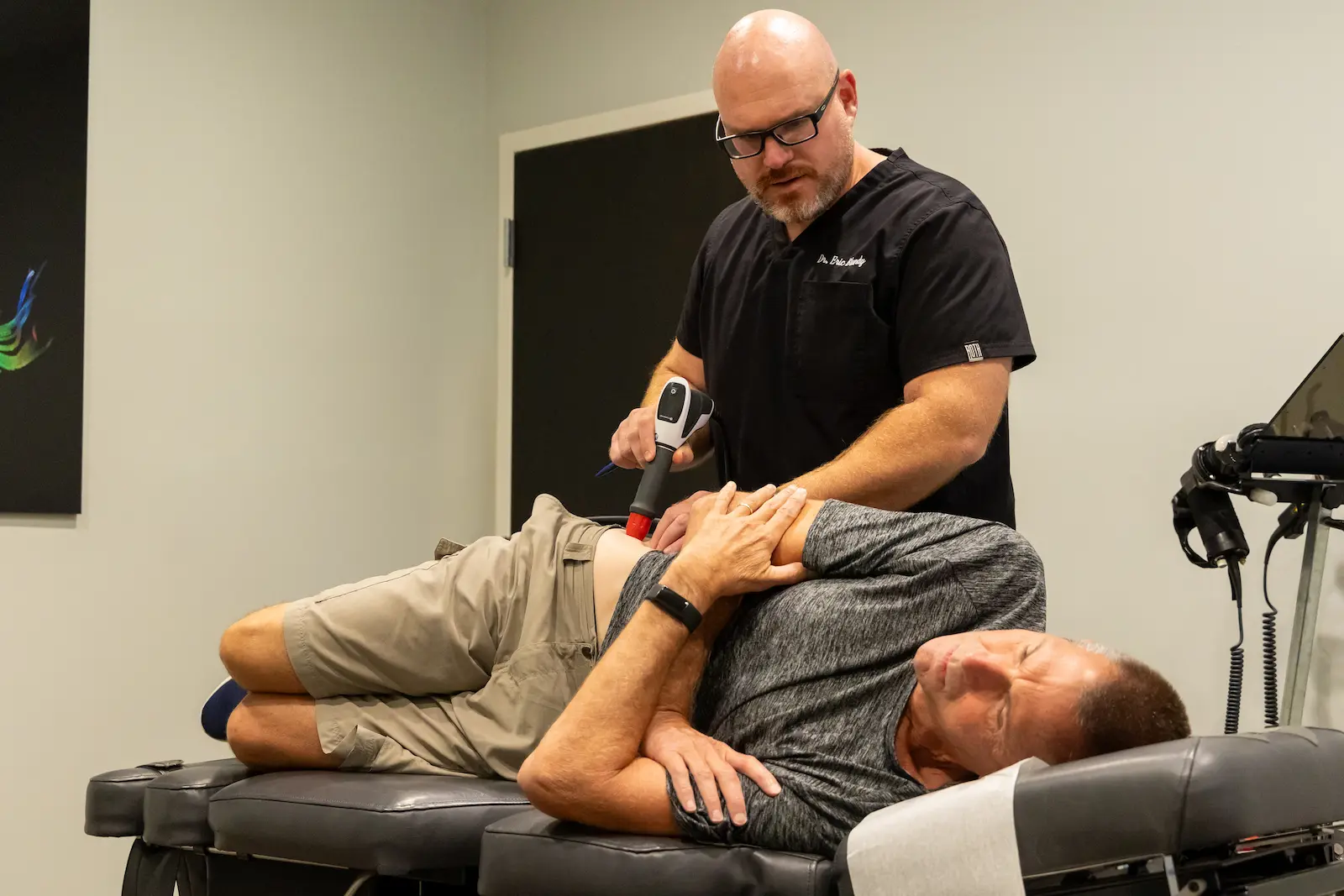 A male practitioner uses a handheld medical device on a male patient's lower back, who is lying on his side on a chiropractic table.