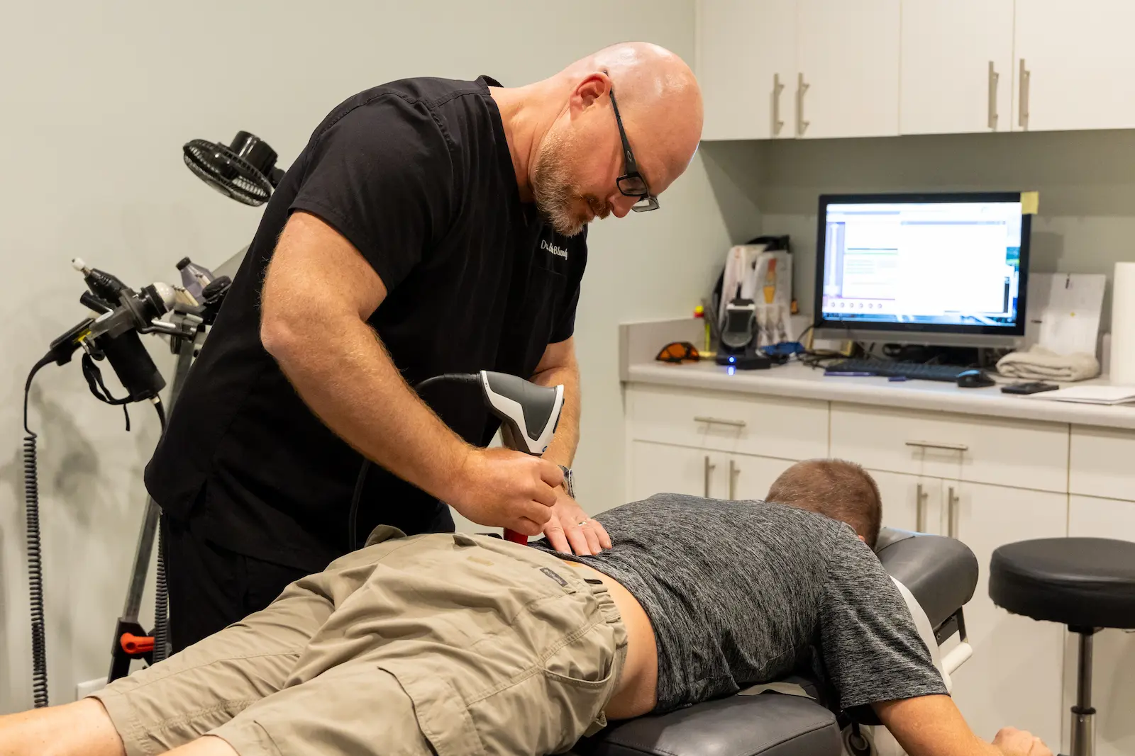 A practitioner in black scrubs uses a handheld device on a patient's lower back as the patient lies face down on a treatment table in a clinic.