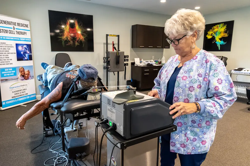 A patient lies face down on a medical table while a female medical professional operates a machine next to them in a clinic setting.