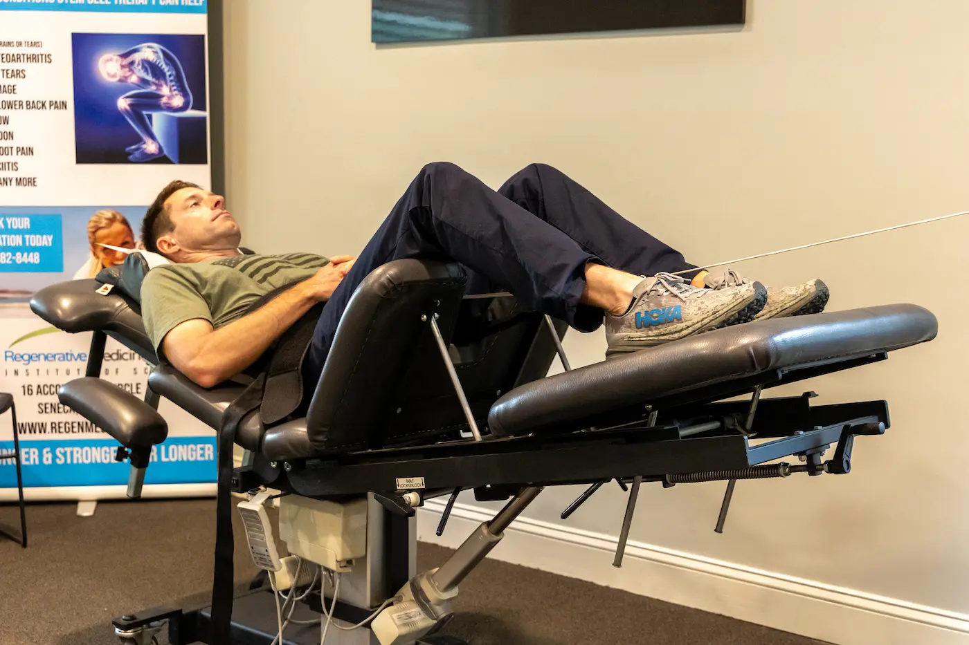 A man lies on his back on a dark grey medical therapy table, with his legs bent and feet elevated. A black strap secures his midsection. A banner for regenerative medicine is in the background.
