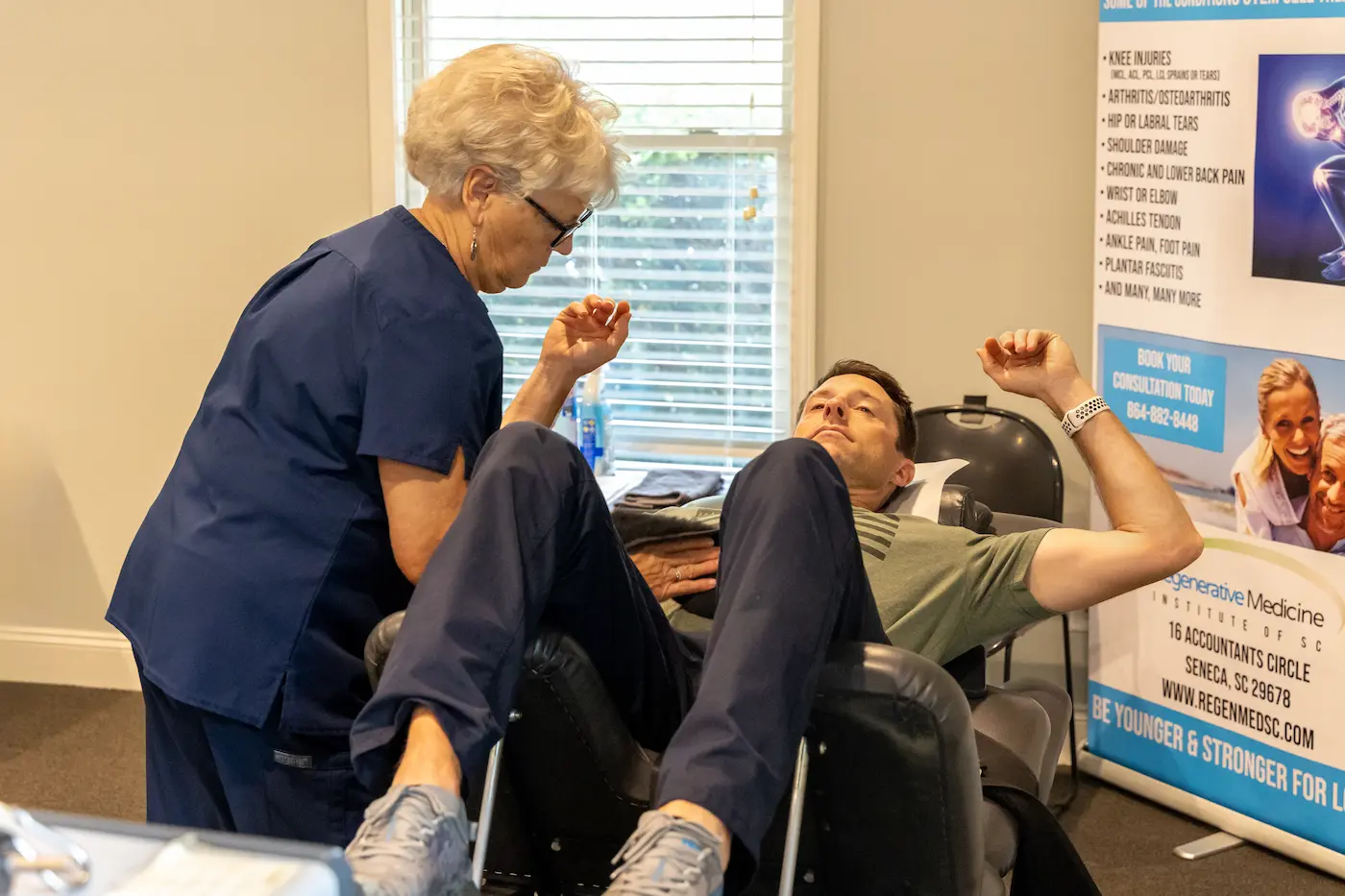 A medical professional in blue scrubs examines a male patient lying in a reclining chair, with a clinic banner in the background.