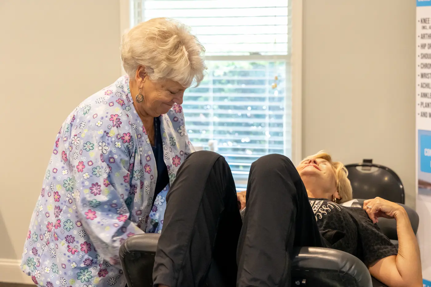 A healthcare worker in a floral scrub top tends to a relaxed patient lying in a black reclining chair.