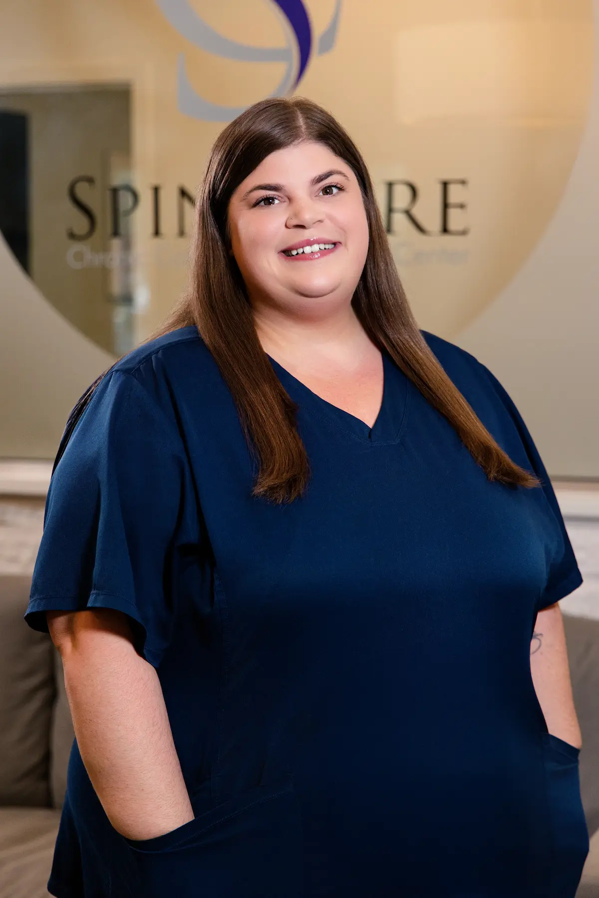 A smiling woman with long brown hair and a dark blue scrub top stands in a professional setting with a blurred logo and text in the background.