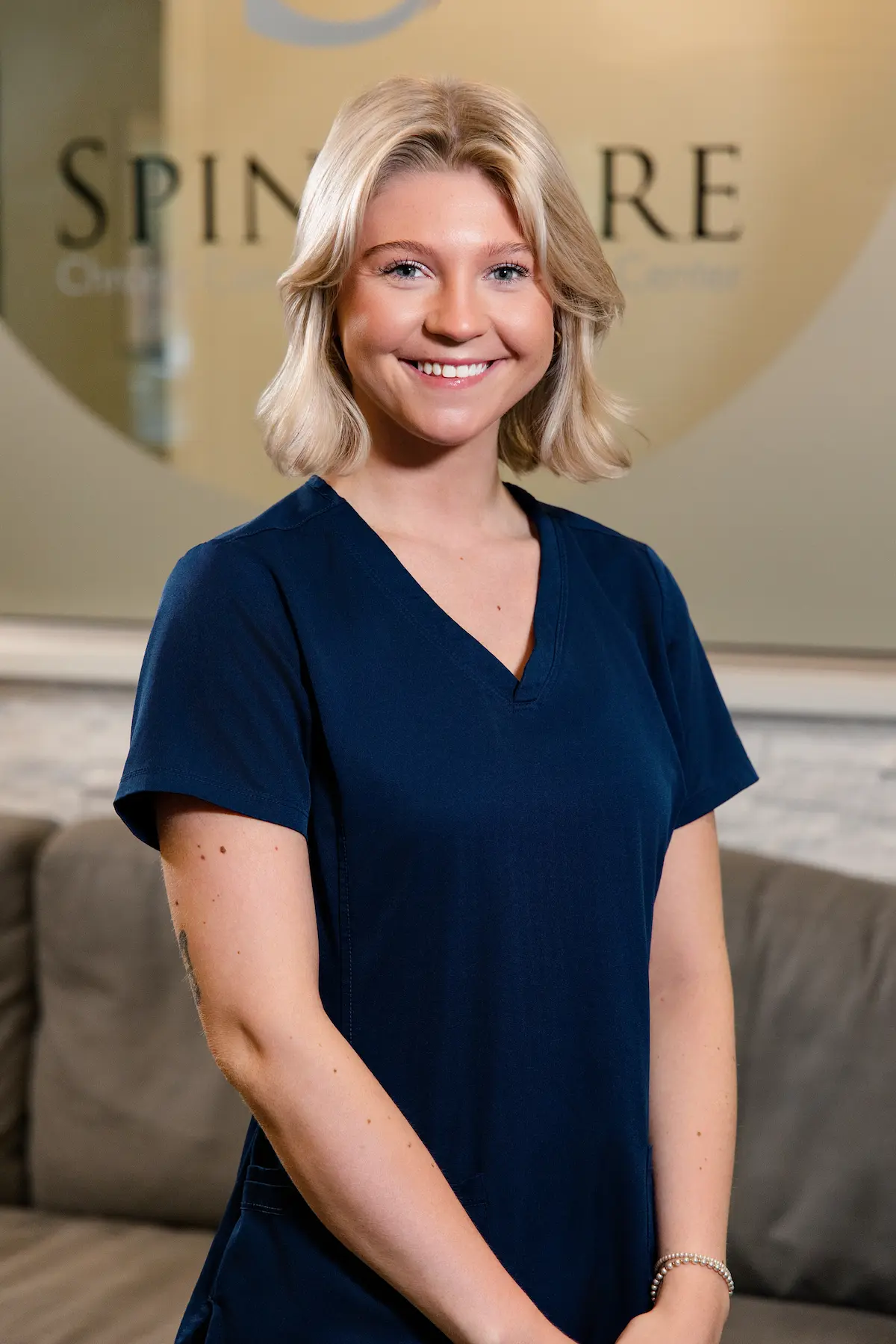 A young woman with blonde hair and blue eyes, wearing dark blue scrubs, smiles at the camera in a professional setting.