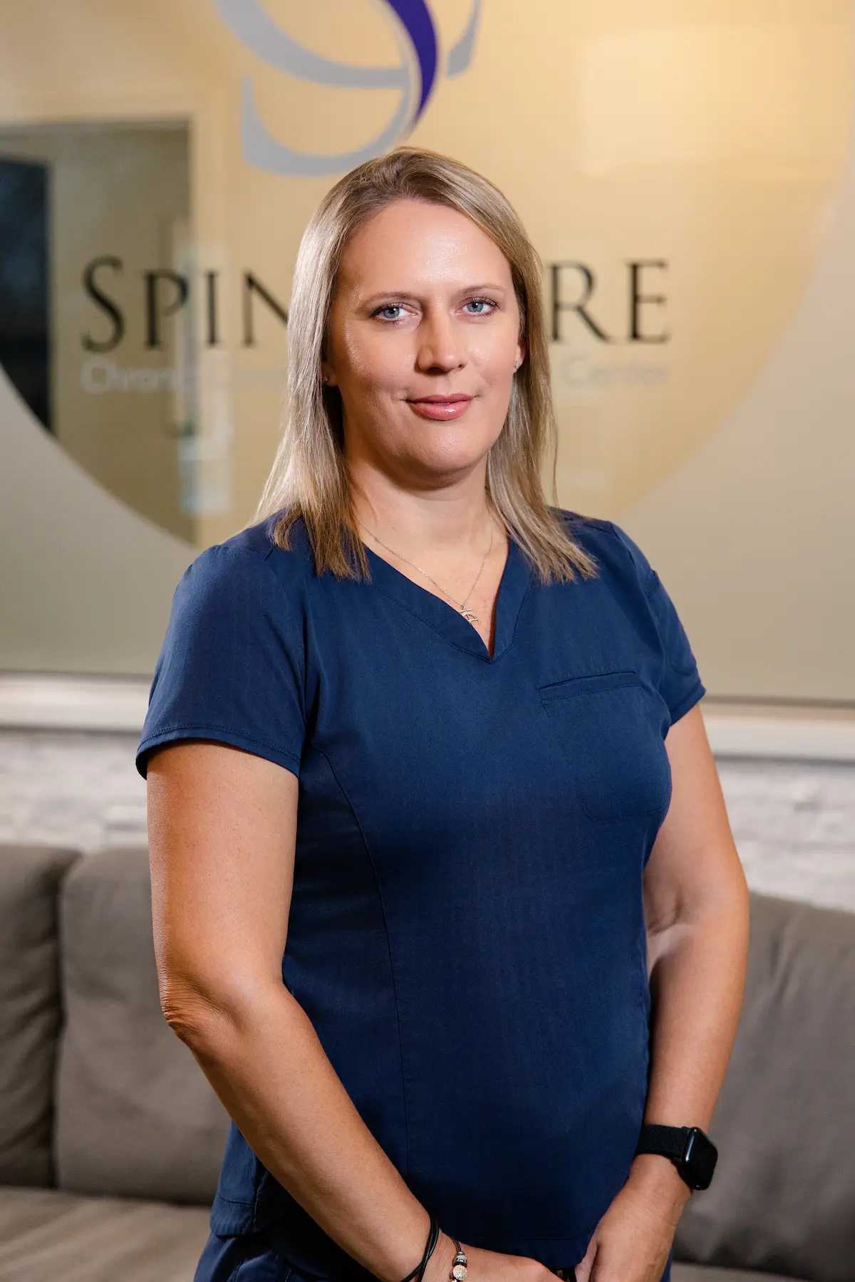 A woman with blonde hair and blue eyes, wearing a navy blue scrub top, stands in a clinic. A blurred logo for 'SPINE CARE Center' is in the background.