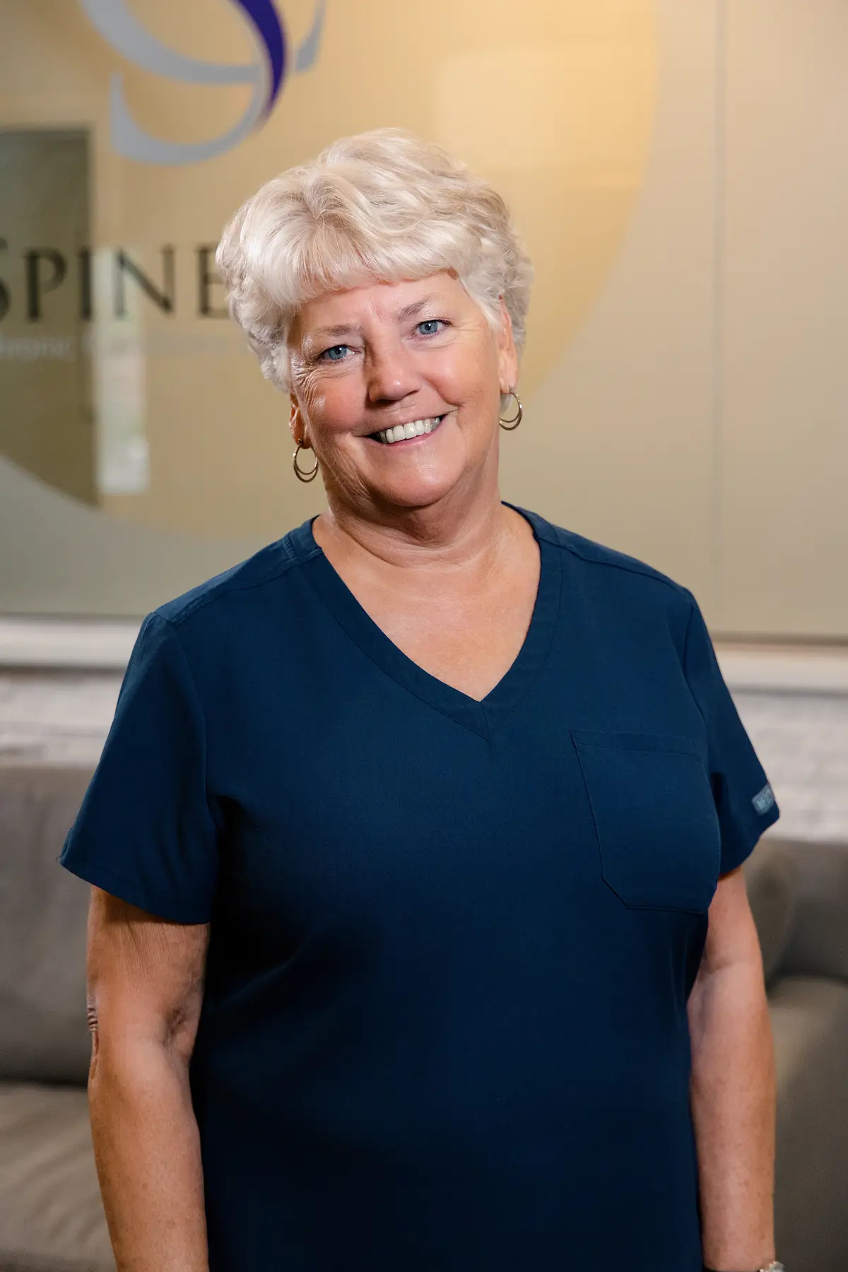 A smiling woman with short blonde hair and blue eyes wearing a dark blue scrub top and hoop earrings.